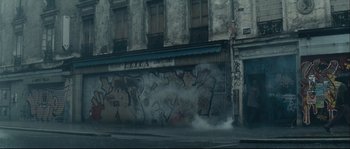 Movie still from “Empire of the Wolves” (2005), directed by Chris Nahon – Smoke billows out of a building on a street corner; Extreme Wide shot, Low angle