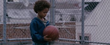 Movie still from “The Pursuit of Happyness” (2006), created by Frank Darabont – A young boy holding a basketball on a chain link fence; Medium shot, Over the shoulder angle