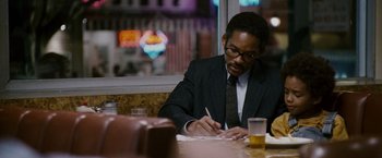 Movie still from “The Pursuit of Happyness” (2006), created by Frank Darabont – A man sitting at a table writing on a piece of paper; Medium shot, Over the shoulder angle