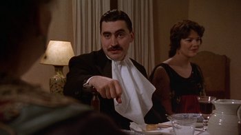 Movie still from “Enchanted April” (1991), directed by Mike Newell – A man in a suit and tie holding a napkin over his face; Medium shot, Over the shoulder angle