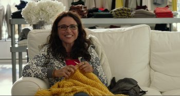 Movie still from “Enough Said” (2013), directed by Nicole Holofcener – A woman sitting on a couch knitting a yellow blanket; Medium shot, Over the shoulder angle