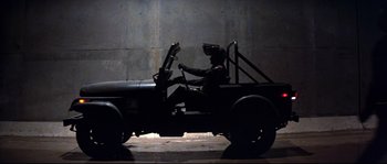 Movie still from “Escape from New York” (1981), directed by John Carpenter – A man in a black suit sitting in the driver's seat of an army jeep; Wide shot, Low angle