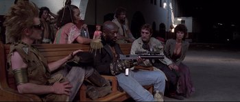 Movie still from “Escape from New York” (1981), directed by John Carpenter – A group of people sitting on a bench playing a guitar; Wide shot, Low angle
