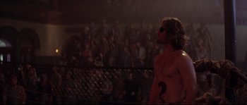 Movie still from “Escape from New York” (1981), directed by John Carpenter – A shirtless man standing in front of an audience; Medium shot, Over the shoulder angle