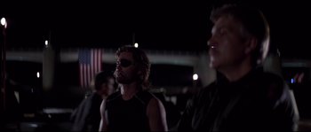 Movie still from “Escape from New York” (1981), directed by John Carpenter – A man in a black shirt and a flag in the background; Medium shot, Over the shoulder angle