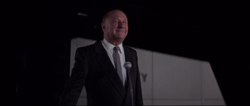 Movie still from “Escape from New York” (1981), directed by John Carpenter – A man in a suit and tie standing in front of a microphone; Medium shot, Low angle