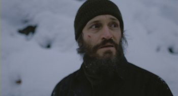 Movie still from “Essential Killing” (2010), directed by Jerzy Skolimowski – A man with a beard wearing a black jacket and a black hat; Close Up shot, Over the shoulder angle