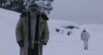Movie still from “Essential Killing” (2010), directed by Jerzy Skolimowski – An older man in a winter coat and hat is holding a stick; Medium shot, High angle