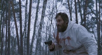 Movie still from “Essential Killing” (2010), directed by Jerzy Skolimowski – A man with a gun in the woods with trees in the background; Medium shot, Low angle