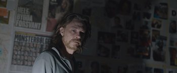 Movie still from “The Quake” (2018), directed by John Andreas Andersen – A man with long hair and a goatee is standing in front of a bunch of pictures; Close Up shot, Over the shoulder angle