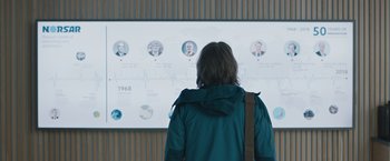 Movie still from “The Quake” (2018), directed by John Andreas Andersen – A woman in a blue jacket is looking at a large screen; Medium shot, Over the shoulder angle