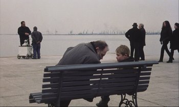 Movie still from “Eternity and a Day” (1998), directed by Theodoros Angelopoulos – A man sitting on top of a wooden bench next to a boy; Wide shot, Over the shoulder angle