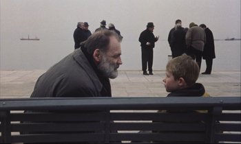 Movie still from “Eternity and a Day” (1998), directed by Theodoros Angelopoulos – A man sitting on top of a park bench next to a boy; Medium shot, Over the shoulder angle