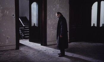 Movie still from “Eternity and a Day” (1998), directed by Theodoros Angelopoulos – A man standing in a dark room with his head turned to the side; Wide shot, Low angle