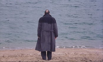Movie still from “Eternity and a Day” (1998), directed by Theodoros Angelopoulos – A man in a long coat standing on the beach; Extreme Wide shot, High angle