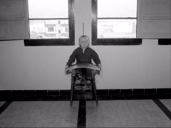 Movie still from “Even Dwarfs Started Small” (1970), directed by Werner Herzog – An old photo of a man sitting in a chair in front of a window; Wide shot, High angle