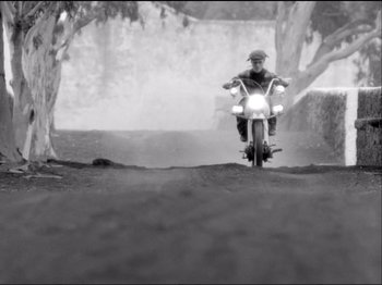 Movie still from “Even Dwarfs Started Small” (1970), directed by Werner Herzog – A man riding a motorcycle down a dirt road; Wide shot, Low angle