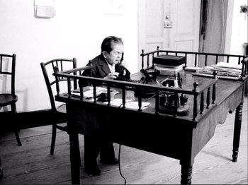 Movie still from “Even Dwarfs Started Small” (1970), directed by Werner Herzog – A man sitting at a table with a pen and paper; Wide shot, High angle
