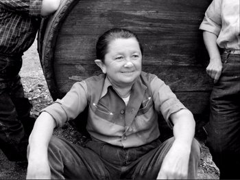 Movie still from “Even Dwarfs Started Small” (1970), directed by Werner Herzog – A woman sitting in front of a wooden barrel; Medium shot, High angle