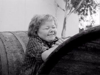 Movie still from “Even Dwarfs Started Small” (1970), directed by Werner Herzog – A young boy is smiling while leaning on a barrel; Medium shot, Low angle