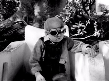 Movie still from “Even Dwarfs Started Small” (1970), directed by Werner Herzog – A little boy sitting in a chair wearing a hat and glasses; Medium shot, High angle