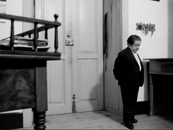 Movie still from “Even Dwarfs Started Small” (1970), directed by Werner Herzog – A little boy standing in front of an open door; Wide shot, Low angle