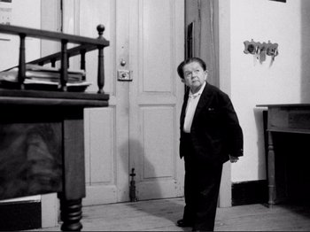 Movie still from “Even Dwarfs Started Small” (1970), directed by Werner Herzog – An older man standing in front of an open door; Medium shot, Low angle