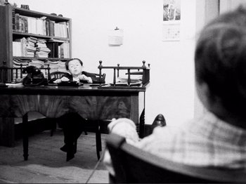 Movie still from “Even Dwarfs Started Small” (1970), directed by Werner Herzog – A man sitting at a table in front of a book shelf; Medium shot, Over the shoulder angle