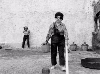 Movie still from “Even Dwarfs Started Small” (1970), directed by Werner Herzog – A young boy wearing sunglasses is holding a stick; Wide shot, Low angle