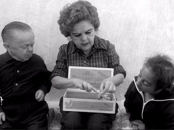 Movie still from “Even Dwarfs Started Small” (1970), directed by Werner Herzog – An old photo of a woman opening a box with two children; Medium shot, High angle