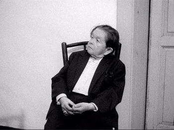 Movie still from “Even Dwarfs Started Small” (1970), directed by Werner Herzog – An older woman sitting in a chair in front of a white wall; Medium shot, High angle