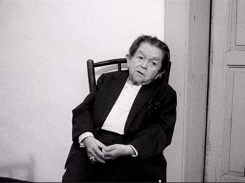 Movie still from “Even Dwarfs Started Small” (1970), directed by Werner Herzog – An older woman sitting in a chair wearing a suit; Medium shot, High angle