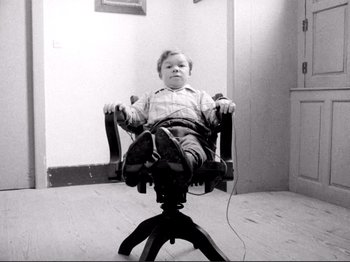 Movie still from “Even Dwarfs Started Small” (1970), directed by Werner Herzog – A little boy sitting in a chair in a room; Wide shot, High angle