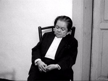Movie still from “Even Dwarfs Started Small” (1970), directed by Werner Herzog – An older woman sitting in a chair with her eyes closed; Medium shot, High angle