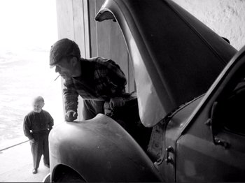 Movie still from “Even Dwarfs Started Small” (1970), directed by Werner Herzog – An old photo of a man looking under the hood of an old car; Medium shot, High angle
