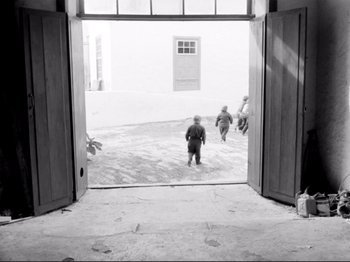 Movie still from “Even Dwarfs Started Small” (1970), directed by Werner Herzog – A black - and - white photo of two children walking through a doorway; Wide shot, High angle