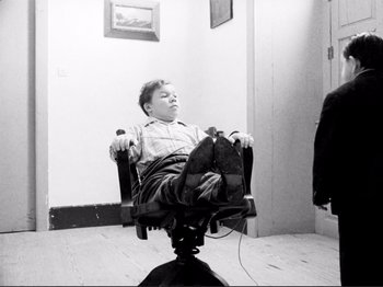 Movie still from “Even Dwarfs Started Small” (1970), directed by Werner Herzog – A boy sitting in a chair with his feet on the floor; Wide shot, High angle