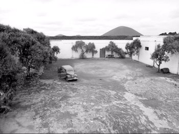 Movie still from “Even Dwarfs Started Small” (1970), directed by Werner Herzog – An old car is parked in an empty parking lot; Extreme Wide shot, High angle
