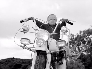 Movie still from “Even Dwarfs Started Small” (1970), directed by Werner Herzog – A man riding a motorcycle on the side of the road; Medium shot, Low angle