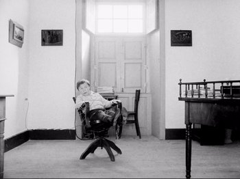 Movie still from “Even Dwarfs Started Small” (1970), directed by Werner Herzog – A man sitting in a chair in front of an open door; Wide shot, High angle