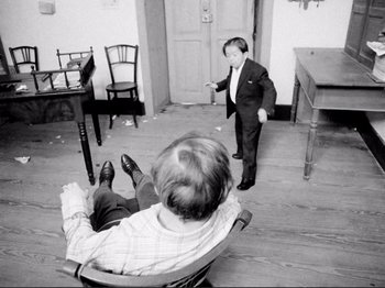Movie still from “Even Dwarfs Started Small” (1970), directed by Werner Herzog – A man in a suit and tie standing in front of another man in a suit and tie in front of a door; Wide shot, High angle