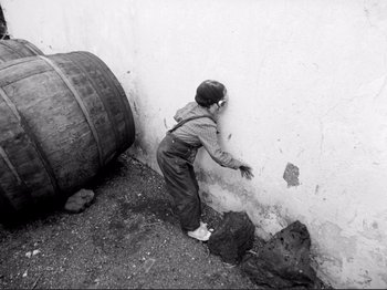 Movie still from “Even Dwarfs Started Small” (1970), directed by Werner Herzog – A young boy in overalls is leaning against a wall; Wide shot, Overhead angle