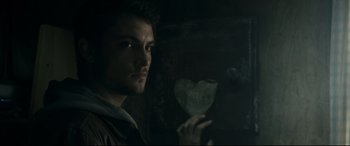 Movie still from “Evil Dead” (2013), directed by Fede Alvarez – A man holding a paper heart in a dimly - lit room; Close Up shot, Over the shoulder angle