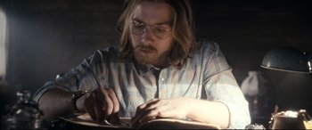 Movie still from “Evil Dead” (2013), directed by Fede Alvarez – A man with long hair and glasses is writing on a piece of paper; Medium shot, Low angle