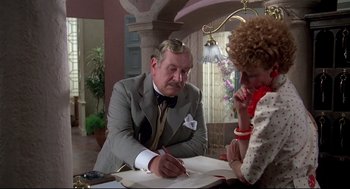 Movie still from “Evil Under the Sun” (1982), directed by Guy Hamilton – An older man in a suit writing on a piece of paper while an older woman looks on; Medium shot, Over the shoulder angle