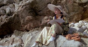 Movie still from “Evil Under the Sun” (1982), directed by Guy Hamilton – A woman sitting on a rock holding a book and wearing a hat; Wide shot, High angle