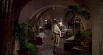 Movie still from “Evil Under the Sun” (1982), directed by Guy Hamilton – An older woman in a white suit is standing in a living room; Wide shot, High angle