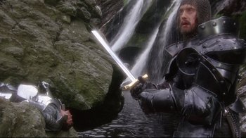 Movie still from “Excalibur” (1981), directed by John Boorman – A man holding a sword in front of a waterfall; Wide shot, Over the shoulder angle