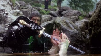 Movie still from “Excalibur” (1981), directed by John Boorman – A person in a body of water holding a sword; Medium shot, Over the shoulder angle