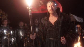Movie still from “Excalibur” (1981), directed by John Boorman – A man with a beard holding a staff in front of a crowd; Medium shot, Over the shoulder angle